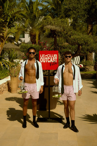 Two shirtless men in pink shorts standing outdoors with a 'Le Club Outcast' sign.
