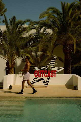 Man walking by a pool with palm trees and 'Outcast' branding in the background